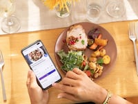 Person using the Weight Watchers app on a smartphone while eating a balanced meal with roasted chicken, vegetables, and salad at a dining table.