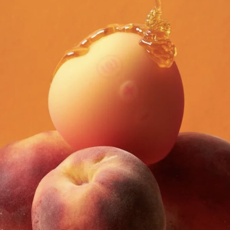 Peach-shaped Womanizer Peach clitoral stimulator in a soft pink-to-yellow gradient, styled on real peaches with honey dripping against a bright orange background.