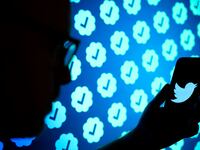 A photo illustrating showing a shadowy figure holding a phone with a Twitter blue bird logo. In the background, there is repetitive imagery of Twitter’s verified badges. Elon Musk recently announced a temporary limit on the number of posts users can read.