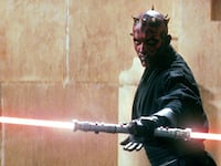 A man with a red and black face holds a lightsaber.