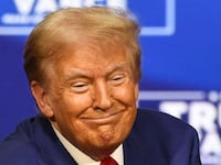 Donald Trump smiles in front of a blue background on stage.