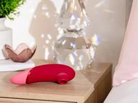 Womanizer Next air suction sex toy in red and pink resting on a bedside table next to decorative crystal lamp and candle, highlighting its sleek whisper-quiet design for Valentine’s Day