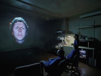 Michael Cera’s disembodied head on a screen, staring straight ahead, as three people sit in armchairs in a dark room below.